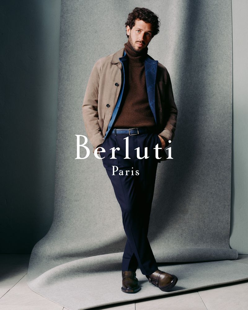 Stylish man in brown turtleneck, jacket, and navy pants poses against a grey backdrop in a fashion advertisement for Berluti Paris.