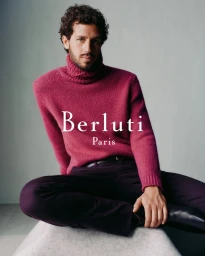 Man wearing a magenta turtleneck sweater and dark trousers seated against a grey studio backdrop for Berluti F/W 2025 Campaign