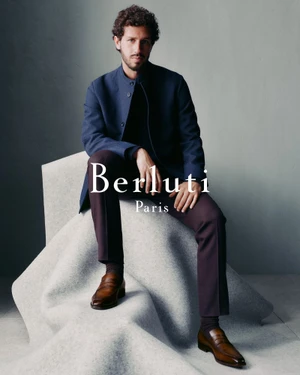 Man seated on a sculpted grey block wearing a blue jacket, dark trousers, and brown leather loafers against a grey background.