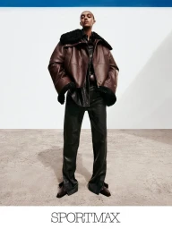 Model wearing a brown shearling-lined leather jacket and black trousers against a neutral minimalist background.