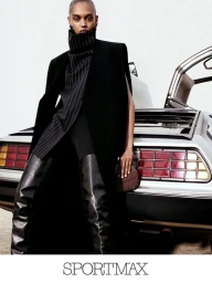 Model standing against a parked car wearing a black coat and tall boots for the Sportmax Fall-Winter 2026 campaign.