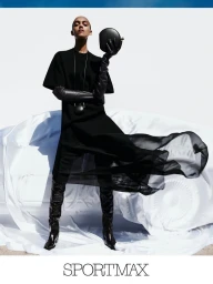 Model in black dress with leather gloves and boots holding a handbag in front of a white draped car for Sportmax