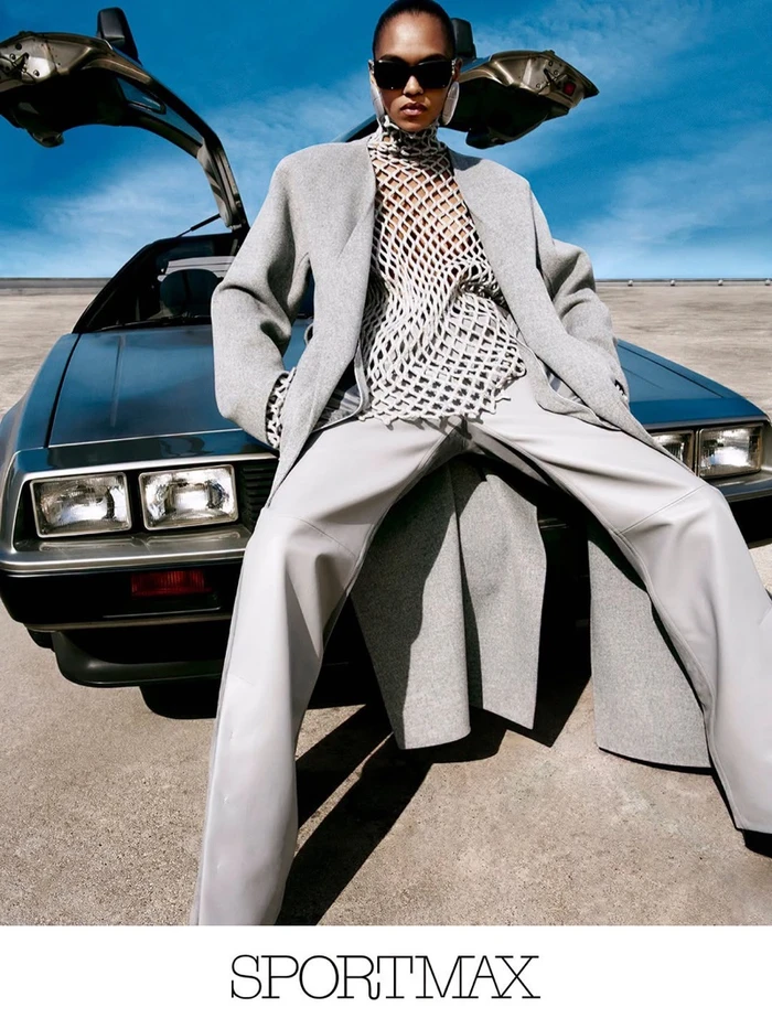 Model in sunglasses and grey coat leaning against a dark car with gull-wing doors open under a clear blue sky