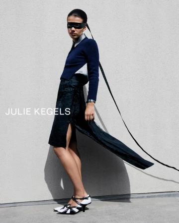 Model in blue sweater and dark patterned skirt wearing eye mask against pale textured wall