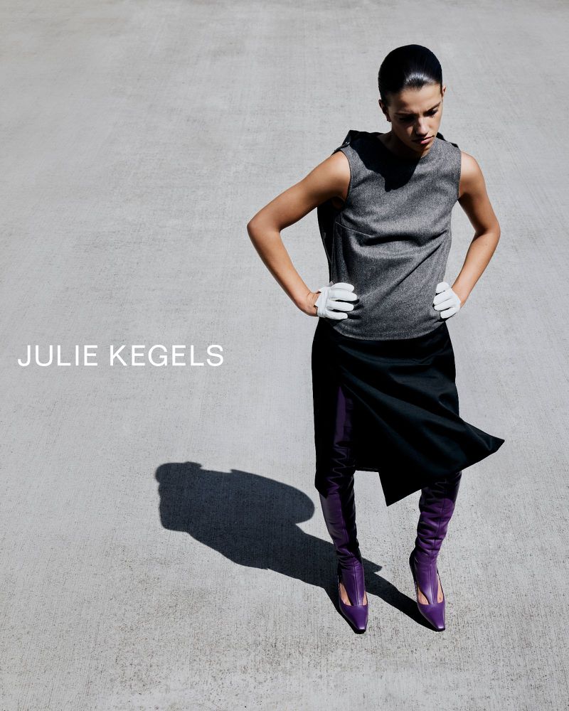 Model wearing a grey sleeveless top, black skirt, white gloves, and purple boots on a concrete surface with visible harsh sunlight shadow