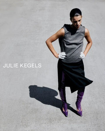 Model wearing a grey sleeveless top, black skirt, white gloves, and purple boots on a concrete surface with visible harsh sunlight shadow