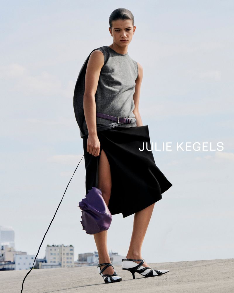 Model wearing a grey top and black skirt with a purple handbag against a sky background