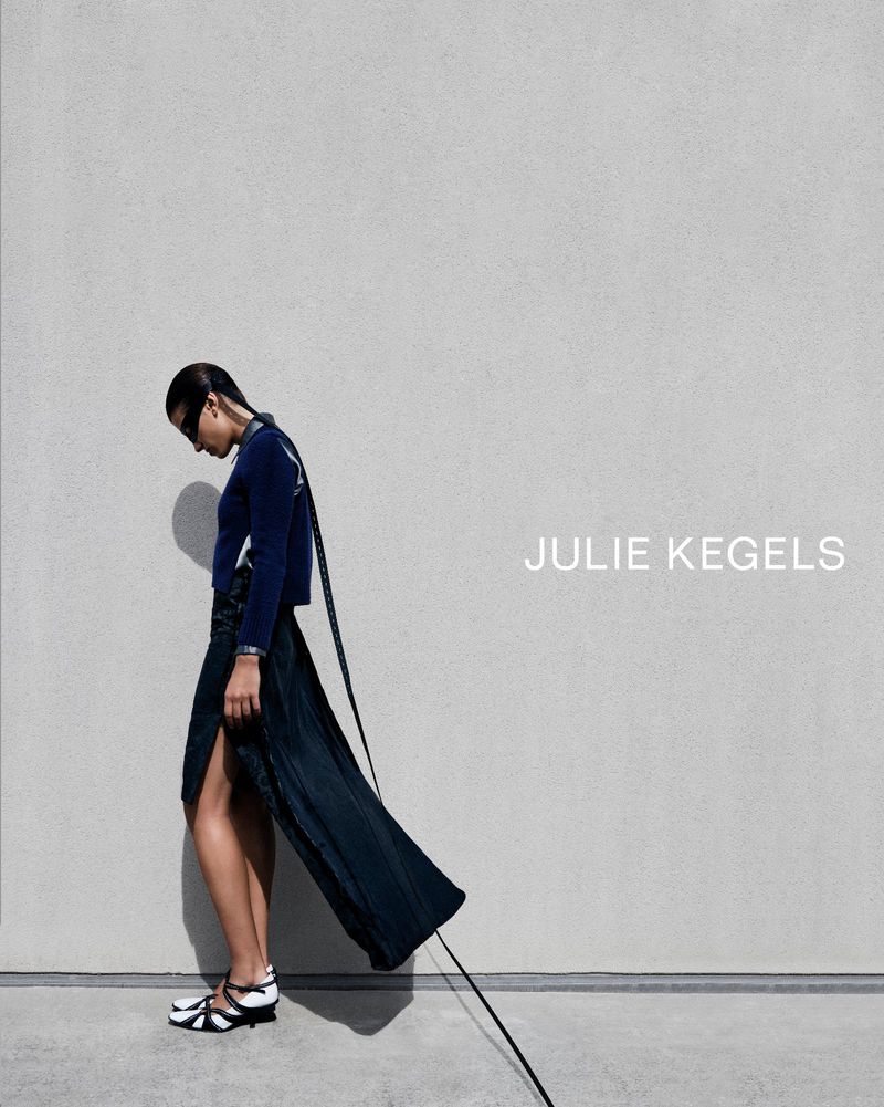 Model in dark blue top and long skirt with strap standing against a concrete wall for Julie Kegels