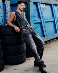 Denis Jovanovic wearing a sleeveless dark vest and charcoal jeans leaning against stacked tires and a blue industrial container.