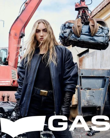 Model in dark leather jacket standing in an industrial scrap yard with a crane lifting a crushed car.