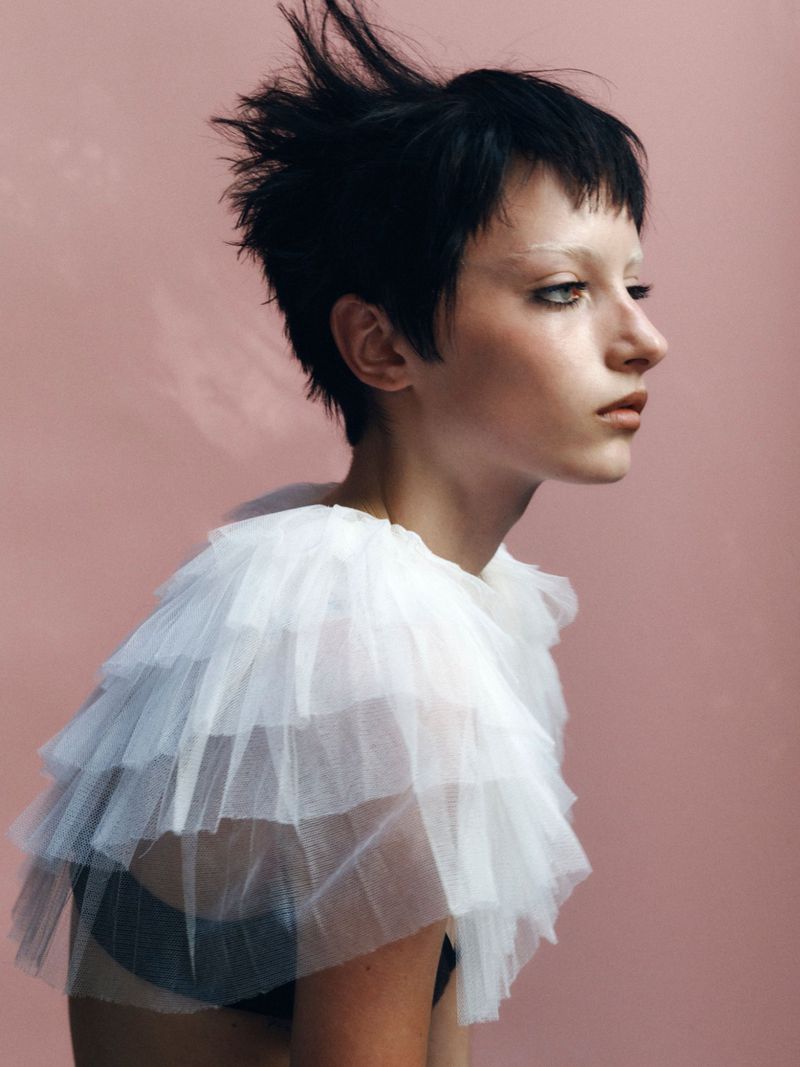 Model with dark spiky hair wearing a white ruffled tulle top against a pink background