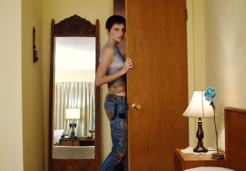 Model Kai Schreiber peeking behind a wooden door in a domestic bedroom, wearing a blue patterned top and patched denim jeans.