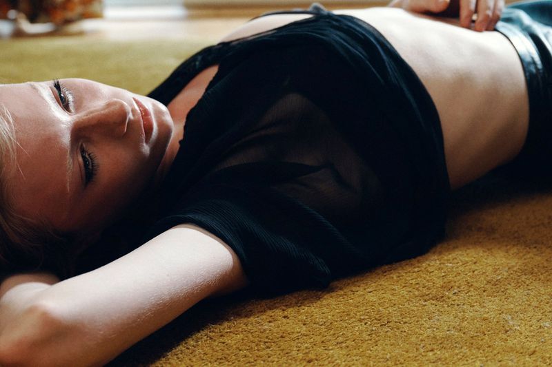 Model lying on a textured yellow carpet wearing a black top and dark trousers in a horizontal fashion photograph