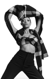 Sophie Wilde in high-contrast black and white, holding a plaid jacket above her head, wearing dark high-waisted trousers against a white backdrop.