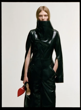 Model in a black leather coat with a high collar holding red-soled black heels against a white background