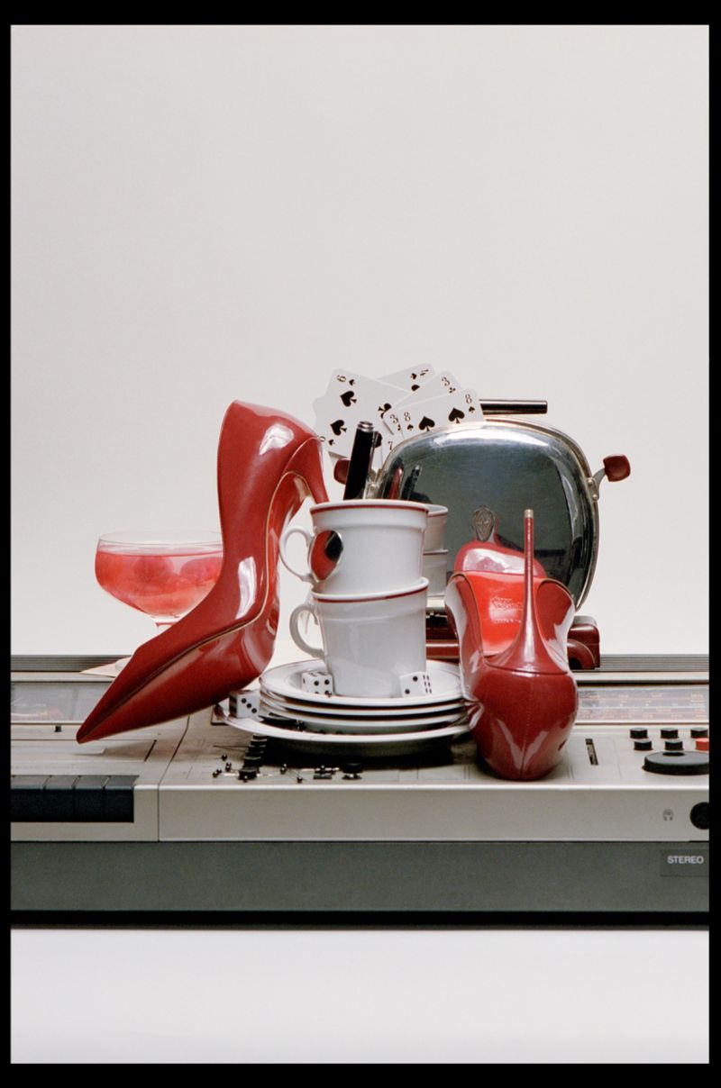 Glossy red high heel shoes arranged on a stereo deck with stacked white cups, playing cards, and a toaster against a plain white backdrop.