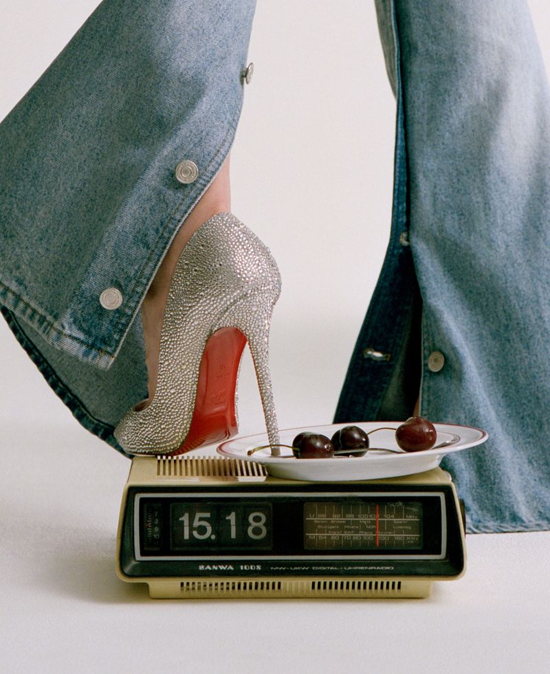 Close-up of a crystal-embellished high heel shoe stepping on a vintage digital clock radio next to a small white plate with cherries.