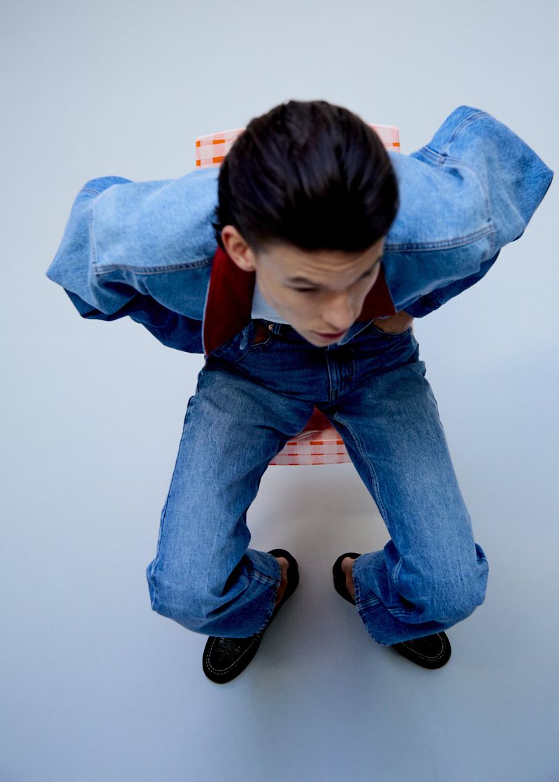 High-angle shot of a person seated wearing a blue denim jacket and matching jeans against a plain backdrop.
