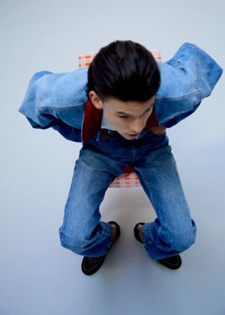 High-angle shot of a person seated wearing a blue denim jacket and matching jeans against a plain backdrop.