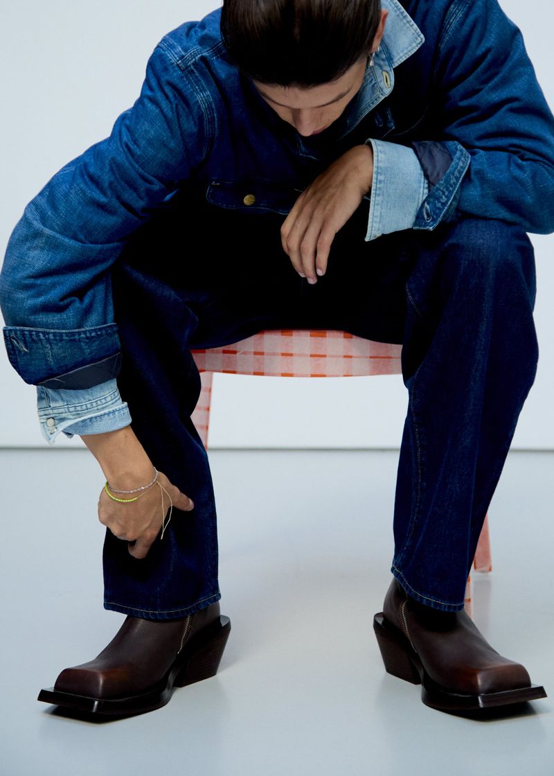 Model seated wearing a blue denim jacket and dark jeans with square-toe brown leather boots against a white studio background.