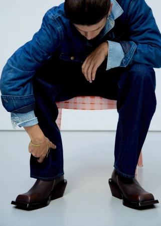Model seated wearing a blue denim jacket and dark jeans with square-toe brown leather boots against a white studio background.