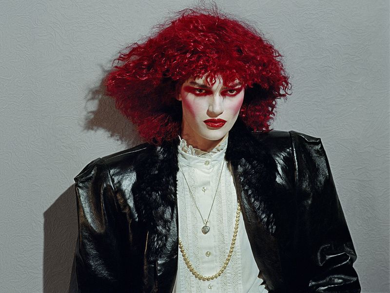 Model with vibrant red curly hair and bold makeup wearing a black jacket and white blouse against a textured grey wall.