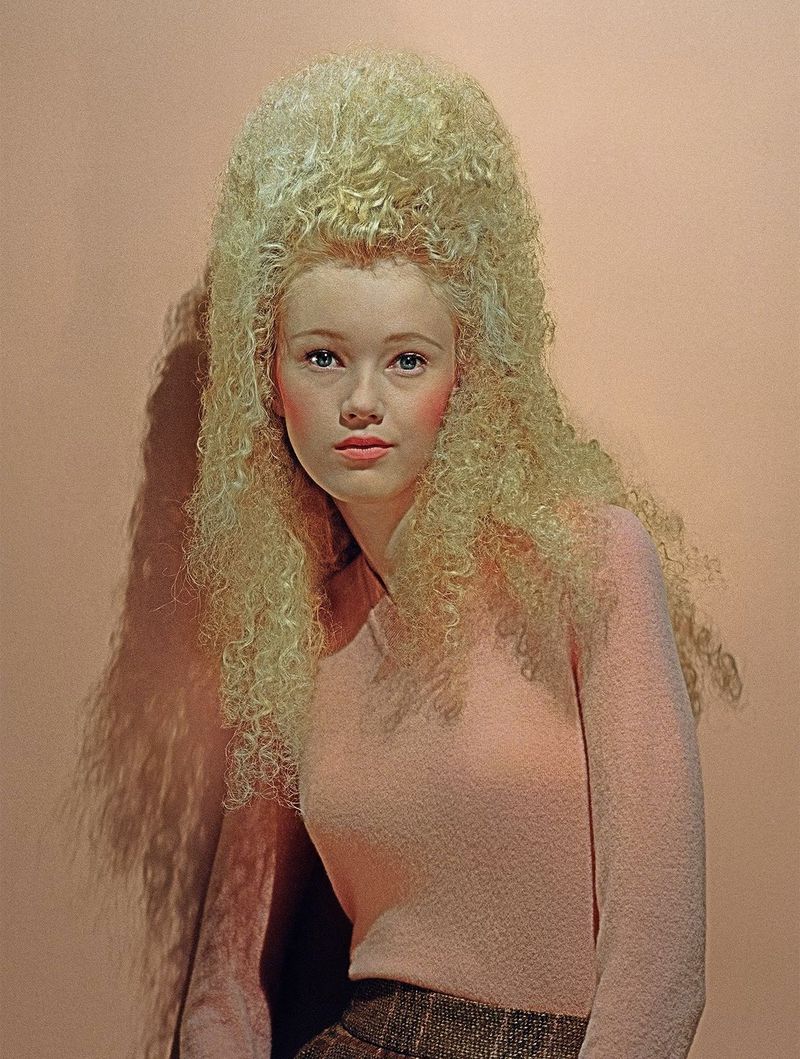 Model with voluminous blonde beehive hair wearing a pink top against a neutral peach backdrop in a New Wave editorial