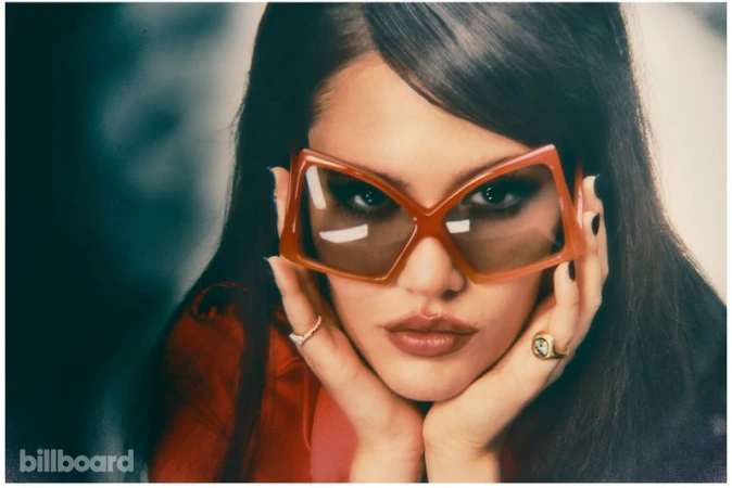 Portrait of Sienna Spiro wearing bold geometric orange sunglasses with dark hair and hands framing her face