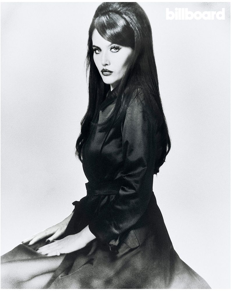 Portrait of Sienna Spiro in a dark belted coat with retro beehive hairstyle against a plain light background for Billboard Magazine.
