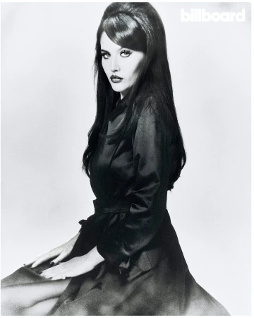 Portrait of Sienna Spiro in a dark belted coat with retro beehive hairstyle against a plain light background for Billboard Magazine.