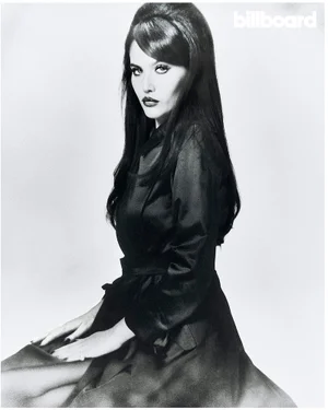 Portrait of Sienna Spiro in a dark belted coat with retro beehive hairstyle against a plain light background for Billboard Magazine.