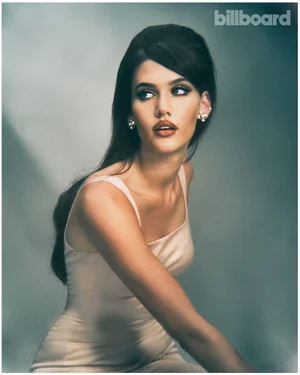 Portrait of Sienna Spiro wearing a cream sleeveless dress and earrings against a textured grey background for Billboard Magazine