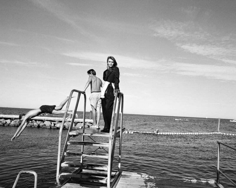 Black and white fashion editorial image showing a model standing on a wooden pier ladder by the sea as another figure dives into the water.