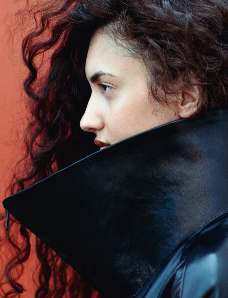 Close-up profile of a woman with dark curly hair wearing a high-collared black leather jacket against a terracotta background.
