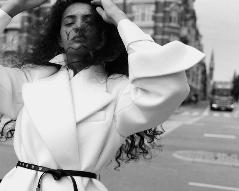 Model with dark curly hair wearing a white coat and thin black belt on a city street