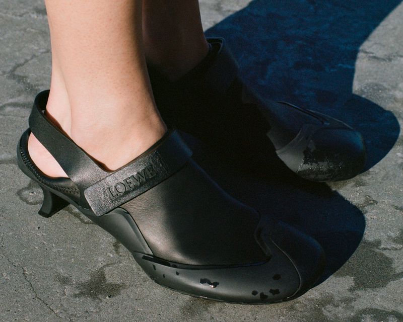Close-up of feet wearing black leather kitten-heel slingback shoes outdoors on a textured grey ground.