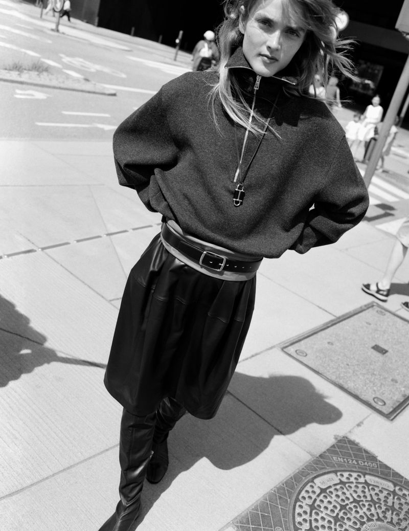 Model standing on a paved city street wearing a zip-up sweater, leather skirt, belt, and high boots.