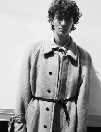 Monochrome portrait of a young man with curly hair wearing a textured belted coat and a patterned neck scarf.