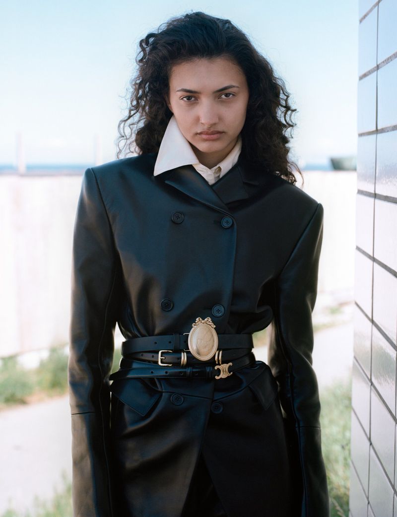 Model with long dark curly hair wearing a black leather jacket and white collar shirt standing outdoors