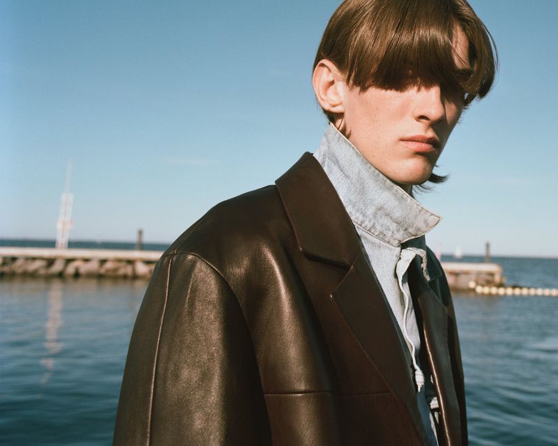 Close-up portrait of a male model wearing a dark leather jacket and denim shirt outdoors by the water under a clear blue sky