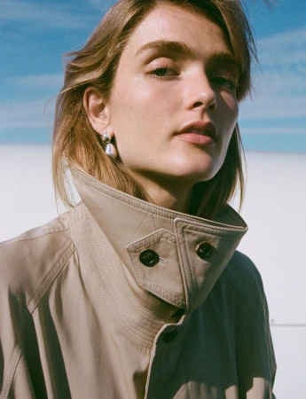 Close-up portrait of a model wearing a high-collared beige jacket and silver drop earrings against a clear sky background