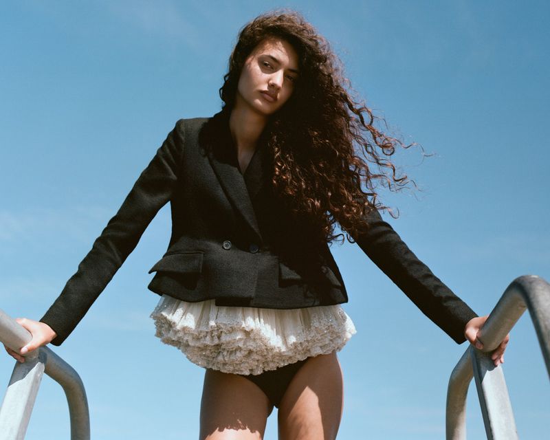 Model with long curly hair wearing a black blazer and white ruffled skirt posing outdoors against a clear blue sky