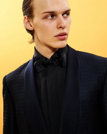 Close-up portrait of a male model in a dark textured blazer and bow tie against a yellow background