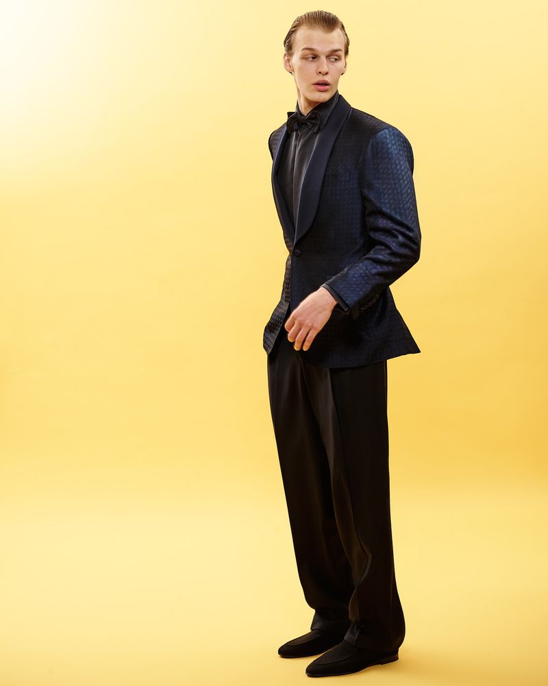 Model in a dark patterned tuxedo jacket and black trousers against a warm yellow backdrop for Canali