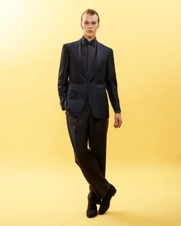 Full-length portrait of a male model wearing a dark tuxedo with a patterned jacket against a warm yellow background