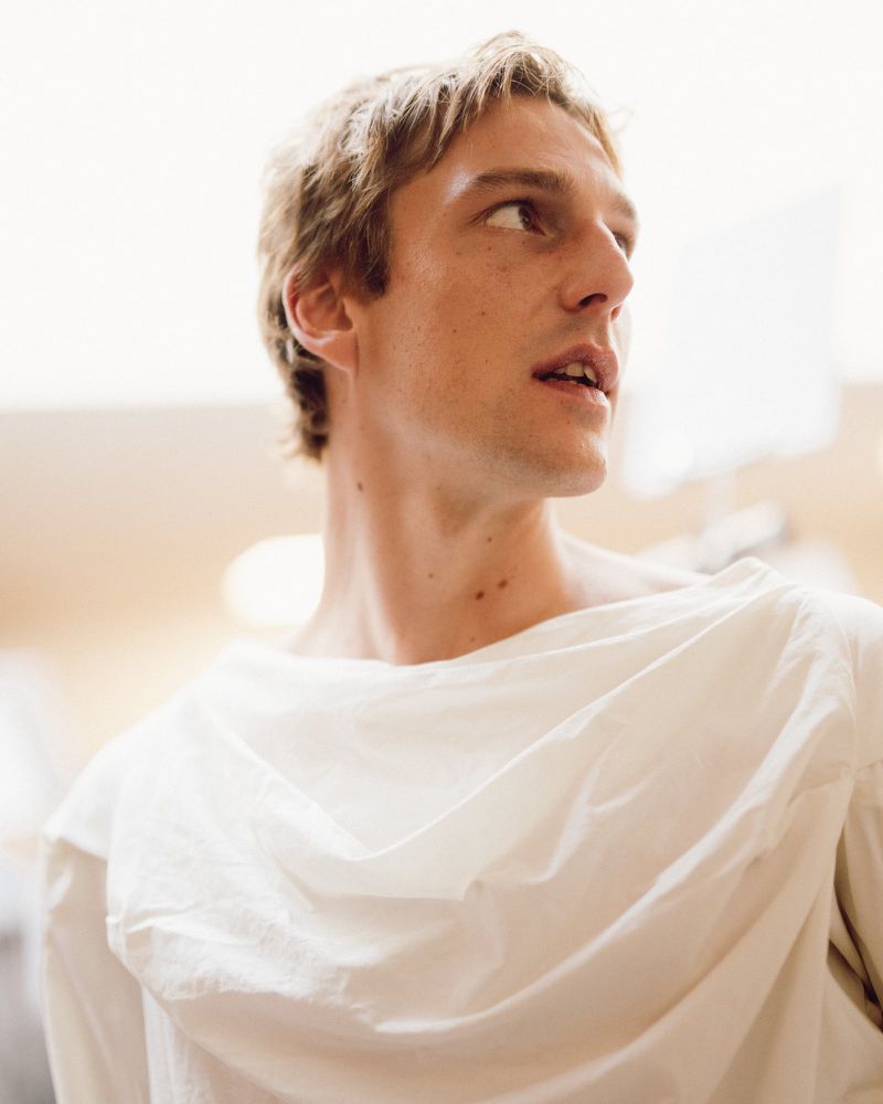 Close-up portrait of a male model wearing a draped white top backstage at Vivienne Westwood S/S 27 captured by Giacomo Cosua.