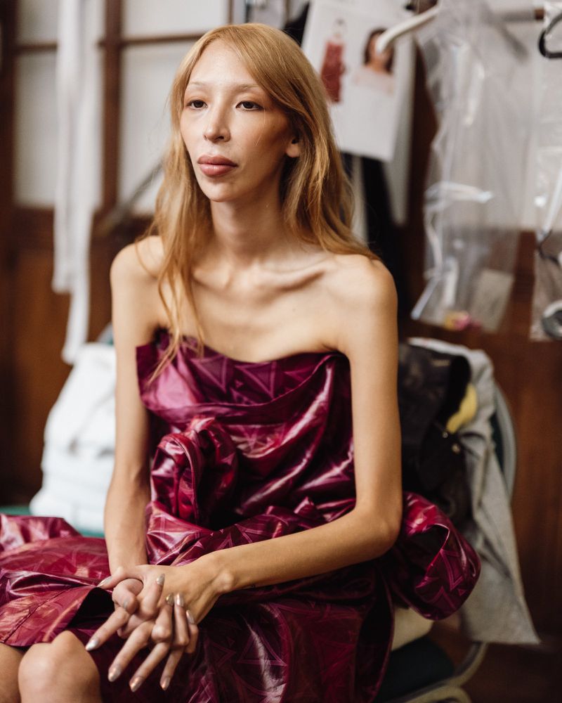 Model wearing a glossy plum strapless dress seated backstage during Vivienne Westwood fashion collection