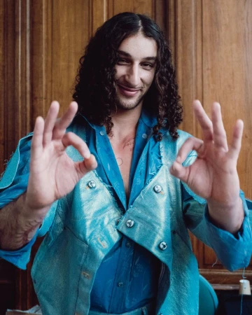 Man with long dark curly hair wearing a blue shirt and jacket gesturing with both hands against a wood-paneled wall