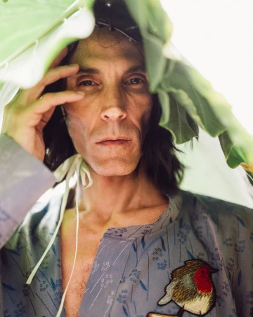 Close-up portrait of a man holding a large green leaf over his head, wearing a patterned grey shirt with an embroidered bird patch.