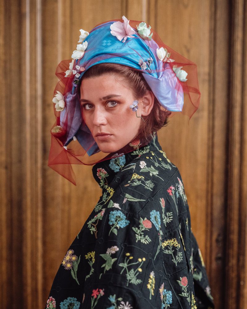 Model wearing a colourful floral headwrap and embroidered black jacket looking over shoulder against wood paneling